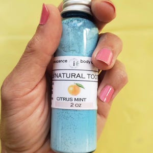 May include: A blue bottle of "SNATURAL TOOTH" citrus mint toothpaste, 57g, held in a hand. The label features an orange graphic. The text "innocence body care" is at the top. The bottle is labeled "Handcrafted", "Small Batches", and "AMAZING taste!"