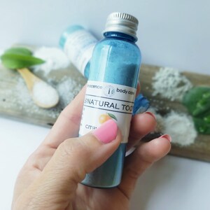 May include: A blue glass bottle with a silver cap, containing blue powder, labelled "i body care SNATURAL TOO". The bottle is held in a hand. Other ingredients and a wooden spoon are in the background.