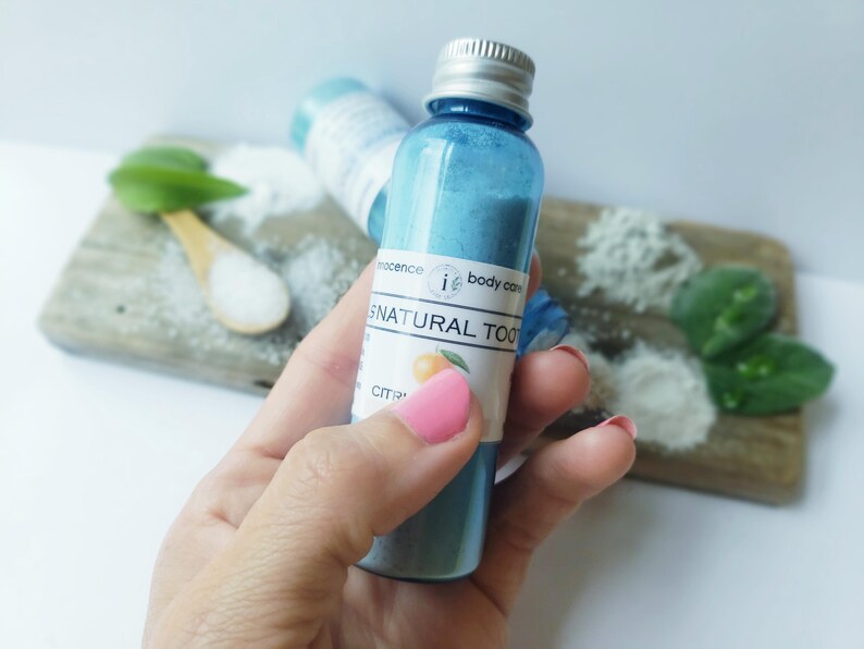 May include: A blue glass bottle with a silver cap, containing blue powder, labelled "i body care SNATURAL TOO". The bottle is held in a hand. Other ingredients and a wooden spoon are in the background.