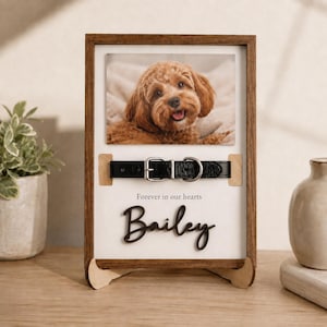 May include: A rectangular wooden memorial frame featuring a photograph of a brown curly-haired dog. Below the photo is a black leather collar with a silver buckle, the text "Forever in our hearts," and the name "Bailey" in black script lettering.