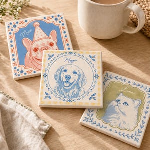 May include: Three square ceramic coasters featuring illustrations of pets. One shows a French bulldog named Max in a party hat, another a golden retriever named Maggie, and the third a cat named Luna. Each coaster has a floral border design.