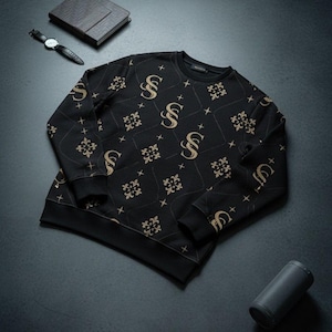 Puede incluir: Sudadera negra de cuello redondo con un patrón dorado repetido de la letra "S" y diseños florales. La sudadera está sobre una superficie oscura, con un reloj, un cuaderno y un altavoz cerca.