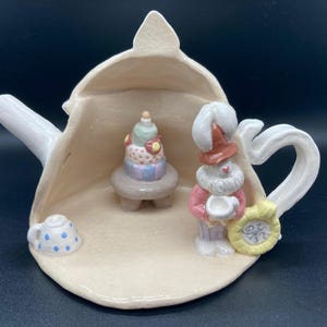 Könnte beinhalten: Eine skurrile Keramik-Teekannen-Figur mit einem Miniaturkuchen auf einem kleinen Tisch und einem Zwerg, der eine Tasse hält. Die Teekanne ist cremefarben, mit Henkel und Ausguss. Der Zwerg trägt einen rot-weißen Hut und einen rosa Mantel.