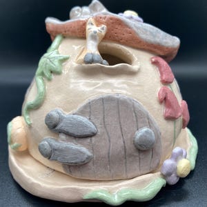 Op de afbeelding: Een grillig keramisch feeënhuis in pastelkleuren. Het huis heeft een lichtbeige lichaam met een grijze deur en handgrepen, een roze en grijs dak en decoratieve bloemen en wijnranken. Een kleine kattenfiguur zit op het dak.