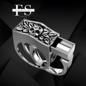 May include: A silver ring with a skull and leaf design. The ring has a rectangular top with a skull and leaf carving. The ring band has a unique design with two circular openings. The ring is from Flacon Sittano Jewelry.