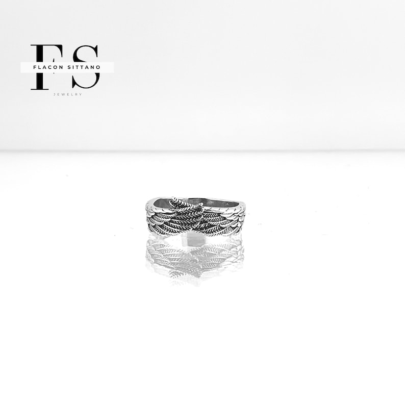 Puede incluir: Un anillo plateado con un dise&ntilde;o detallado de plumas. El anillo est&aacute; centrado y se refleja en la superficie blanca. La marca "Flacon Sittano Jewelry" est&aacute; en la esquina superior izquierda.