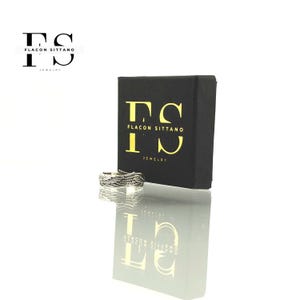 Puede incluir: Un anillo plateado con un dise&ntilde;o tejido se muestra junto a una caja de joyer&iacute;a negra. La caja tiene letras doradas que dicen "Flacon Sittano Jewelry". El anillo se refleja en la superficie brillante de abajo.