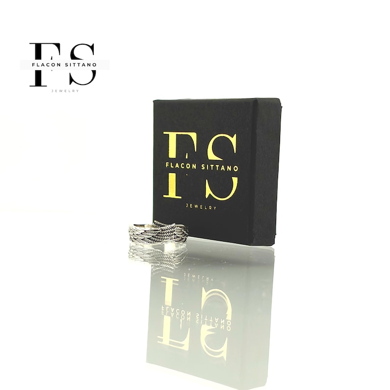 Puede incluir: Un anillo plateado con un dise&ntilde;o tejido se muestra junto a una caja de joyer&iacute;a negra. La caja tiene letras doradas que dicen "Flacon Sittano Jewelry". El anillo se refleja en la superficie brillante de abajo.