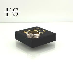 Puede incluir: Un anillo dorado con un dise&ntilde;o detallado de plumas. El anillo se muestra en una caja negra con letras doradas. Joyer&iacute;a de Flacon Sittano.