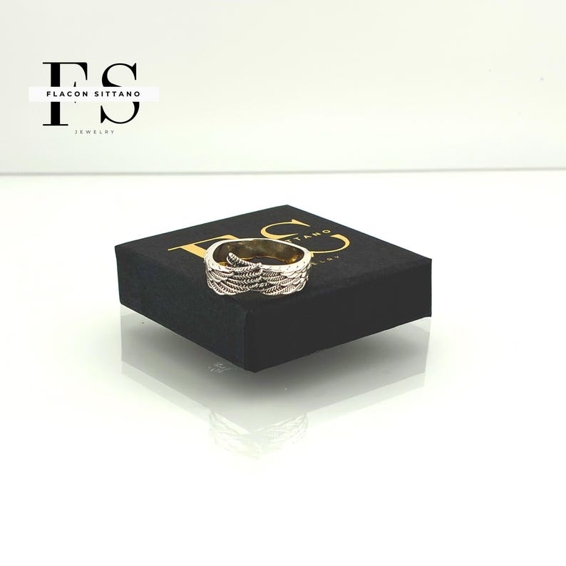 Puede incluir: Un anillo dorado con un dise&ntilde;o detallado de plumas. El anillo se muestra en una caja negra con letras doradas. Joyer&iacute;a de Flacon Sittano.