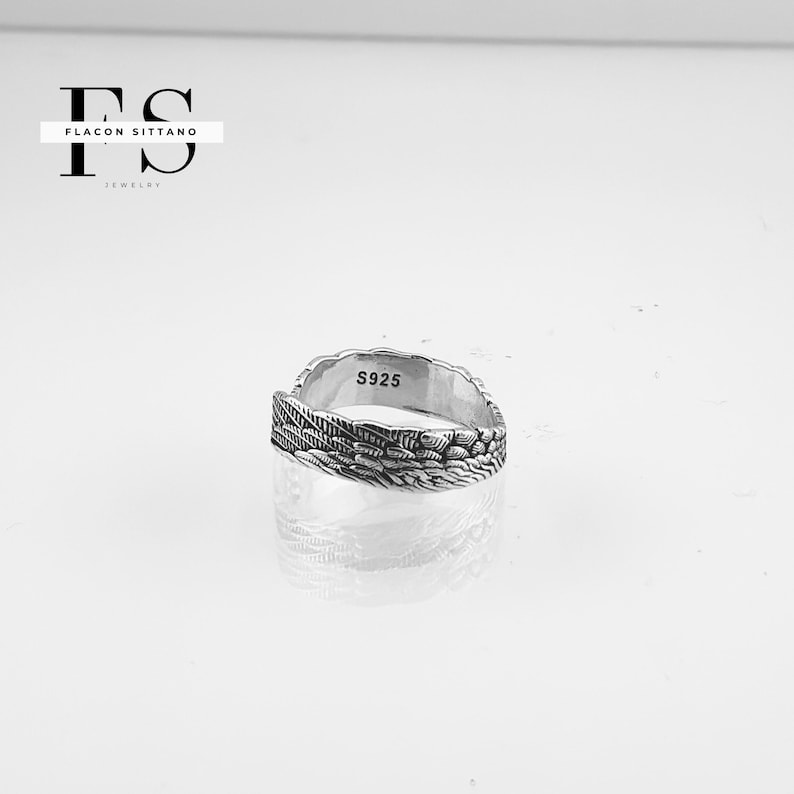 Puede incluir: Un anillo plateado con un dise&ntilde;o detallado de plumas. El anillo est&aacute; marcado con "S925" y se encuentra sobre un fondo blanco. Es una pieza de joyer&iacute;a.