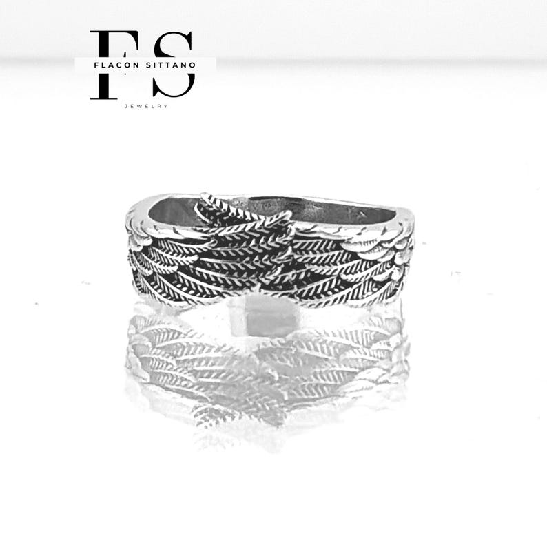 Puede incluir: Un anillo plateado con un dise&ntilde;o detallado de alas. El anillo es muy reflectante, con el dise&ntilde;o de alas envolviendo la banda. La imagen incluye el texto "FLACON SITTANO JEWELRY" en la esquina superior izquierda.