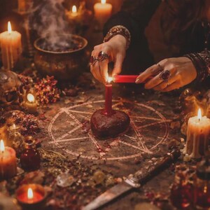 K&ouml;nnte beinhalten: Eine mystische Szene mit Kerzen, einem Pentagramm und einem herzf&ouml;rmigen Objekt. Eine Person z&uuml;ndet eine rote Kerze an, umgeben von anderen brennenden Kerzen, einem Kessel und einem Messer. Die Szene spielt auf einem Tisch mit getrockneten Kr&auml;utern und Blumen.