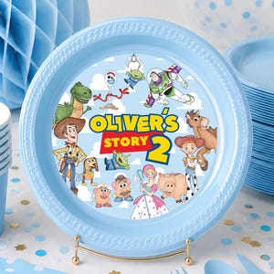 May include: A light blue plastic party plate featuring a custom graphic with the text Oliver's Story 2, surrounded by various animated characters including a cowboy, space ranger, and cowgirl, with a textured rim and a matching set of party supplies.