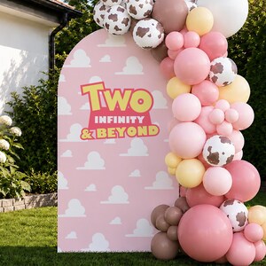 May include: A pink archway with the text "Two Infinity & Beyond" is decorated with a balloon garland. The garland features white, pink, brown, and cow-print balloons. The scene is set outdoors on a green lawn.