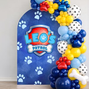 Può includere: Uno sfondo blu con impronte di zampe e la scritta "Leo's Patrol" in un design a scudo, completato da un arco di palloncini blu, gialli, rossi e bianchi. È incluso anche un palloncino a forma di osso.
