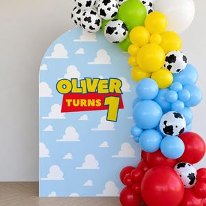 May include: A party decoration featuring a blue arch with white cloud designs and the text "OLIVER TURNS 1". A balloon garland includes black and white cow-print balloons, red, yellow, blue, green, and white balloons.
