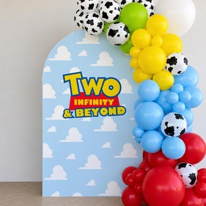 Op de afbeelding: Een blauwe boogvormige achtergrond met witte wolkenontwerpen en de tekst "Two Infinity & Beyond" in geel en rood. Een ballonslinger heeft zwart-witte koeienprint ballonnen, groene, gele, blauwe en rode ballonnen en witte ballonnen.