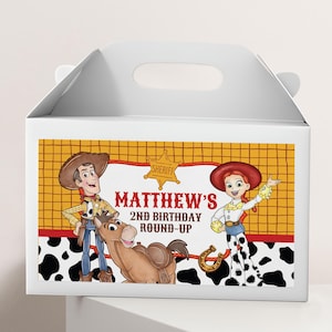 Könnte beinhalten: Eine weiße Pappschachtel mit Tragegriff und einem farbenfrohen Grafikdruck von zwei Zeichentrickfiguren und einem Pferd. Der Text lautet "MATTHEW'S 2ND BIRTHDAY ROUND-UP" mit einem Sheriffstern-Symbol. Das Design zeigt gelbe Holzmaserung und Kuhflecken.
