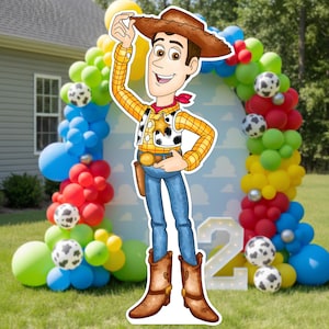May include: A cardboard cutout of the character Woody from Toy Story, wearing a cowboy hat, yellow plaid shirt, and blue jeans. A colourful balloon arch with football accents frames the character, with a large number "2" in white.