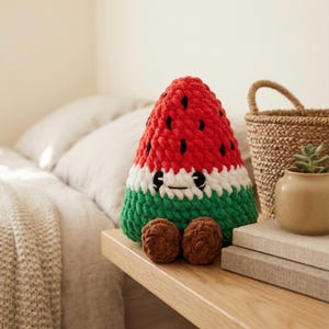 Puede incluir: Una rebanada de sandía de peluche hecha a mano, con una cara sonriente. La parte superior roja tiene detalles de semillas negras, rayas blancas y verdes y pies redondos marrones. El juguete de peluche está sobre una superficie de madera.