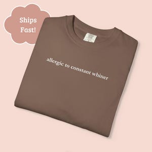May include: A brown t-shirt with the phrase "allergic to constant whiner" printed in white. The shirt is folded and displayed on a light pink background. The shirt has a crew neck and short sleeves.