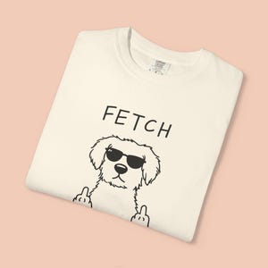 May include: Cream-coloured t-shirt featuring the word "FETCH" above a cartoon dog wearing sunglasses. The dog is giving the middle finger with both paws. The shirt is folded and has a tag in the upper right corner.