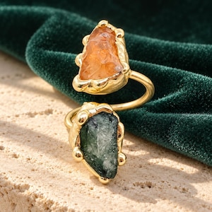 May include: A gold-toned open ring featuring two raw, uncut gemstones. One stone is a translucent orange, while the other is a dark, textured green. The metal band has a textured, organic setting that holds each stone securely in place.