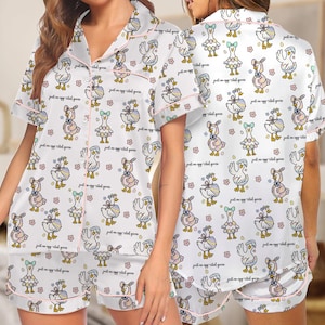 Puede incluir: Conjunto de pijama blanco con un estampado repetido de gansos de dibujos animados con varios atuendos, con ribete rosa. El conjunto incluye una camisa de manga corta con botones y pantalones cortos a juego. La frase "just an egg-cited goose" est&aacute; escrita en la tela.