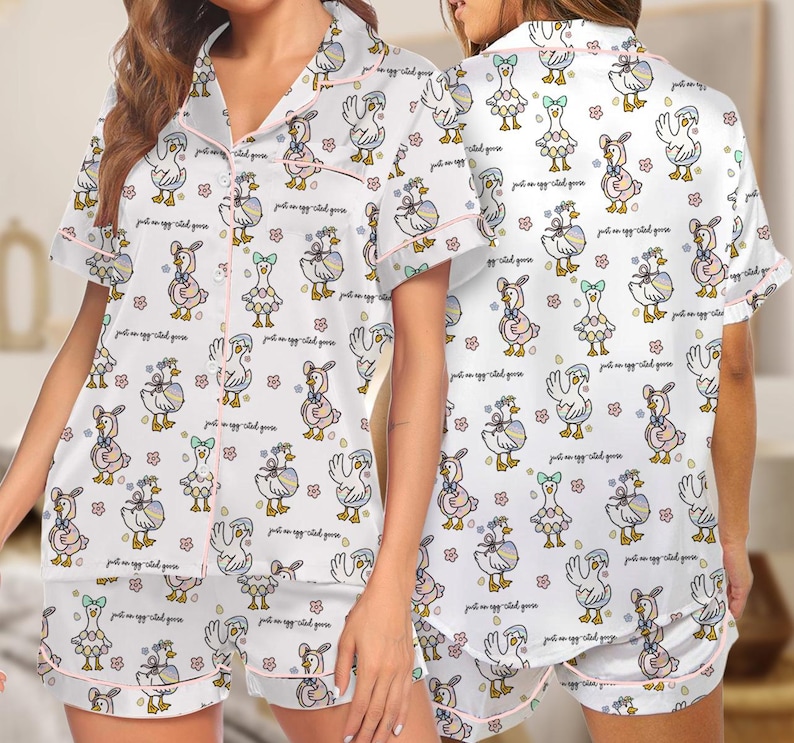 Puede incluir: Conjunto de pijama blanco con un estampado repetido de gansos de dibujos animados con varios atuendos, con ribete rosa. El conjunto incluye una camisa de manga corta con botones y pantalones cortos a juego. La frase "just an egg-cited goose" est&aacute; escrita en la tela.