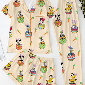 May include: A three-piece pajama set featuring a light beige background with a repeating pattern of Disney characters in Easter egg designs and carrots. The set includes a short-sleeved button-up shirt, shorts, and long pants.