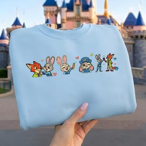 以下が含まれることがあります： Zootopiaの漫画のキャラクターが刺繍された、明るい青色のスウェットシャツ。デザインには、ニック・ワイルド、ジュディ・ホップス、その他のキャラクターが含まれています。スウェットシャツは人が持っています。