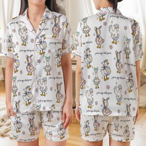 Puede incluir: Conjunto de pijama blanco con un estampado caprichoso de gansos. La camiseta de manga corta y los pantalones cortos presentan un patr&oacute;n repetido de gansos de dibujos animados con diferentes atuendos, con ribete rosa y el texto "just an egg-cited goose".