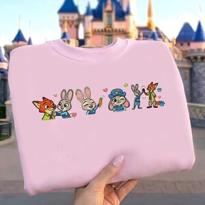 以下が含まれることがあります： 刺繍された漫画のキャラクターが特徴の薄いピンク色のスウェットシャツ。デザインには、キツネ、ウサギ、警察官のウサギが含まれています。キャラクターはさまざまなポーズで、ハートと星があります。スウェットシャツは城の前で持ち上げられています。