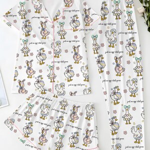 Puede incluir: Conjunto de pijama blanco con estampado de gansos. El conjunto incluye una camisa de manga corta con botones, pantalones cortos y pantalones largos. Cada pieza est&aacute; ribeteada con ribetes rosas y presenta un patr&oacute;n repetido de gansos de dibujos animados y flores. El texto "just an egg cited goose" tambi&eacute;n est&aacute; impreso.