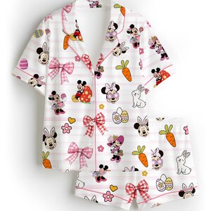 Puede incluir: Conjunto de pijama blanco con estampado de dibujos animados con Minnie Mouse en dise&ntilde;os tem&aacute;ticos de Pascua. El conjunto incluye una camisa con botones y pantalones cortos, ambos adornados con lazos y rayas rosas. Tambi&eacute;n se presentan zanahorias, huevos y flores.