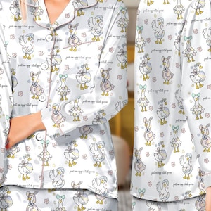 Puede incluir: Conjunto de pijama de sat&eacute;n blanco con un estampado repetido de gansos de dibujos animados con lazos y huevos. El pijama tiene una parte superior abotonada con ribete rosa y pantalones a juego. El texto "just an egg-cited goose" est&aacute; impreso en la tela.