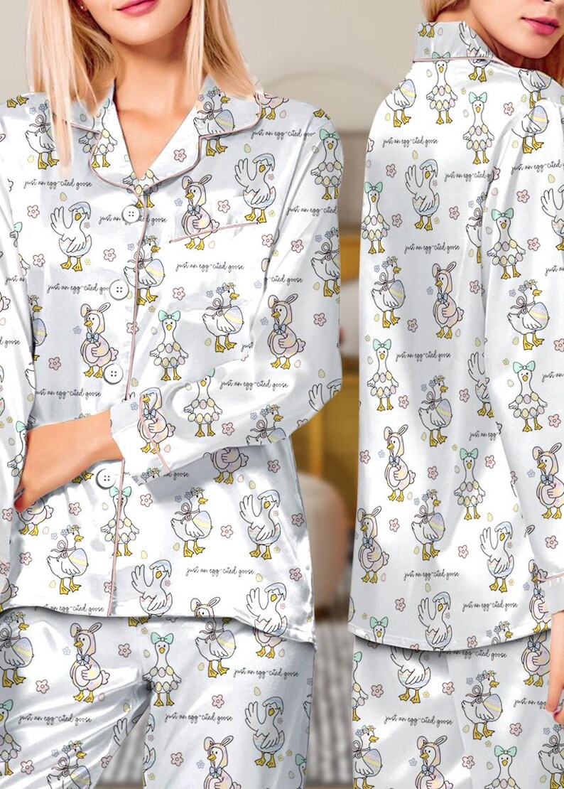 Puede incluir: Conjunto de pijama de sat&eacute;n blanco con un estampado repetido de gansos de dibujos animados con lazos y huevos. El pijama tiene una parte superior abotonada con ribete rosa y pantalones a juego. El texto "just an egg-cited goose" est&aacute; impreso en la tela.