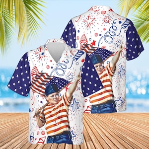 May include: A short-sleeved button-down shirt with a patriotic design. The shirt features a white background with red, white, and blue fireworks and stars. The sleeves are blue with white stars. The shirt has a graphic of a person holding an American flag.