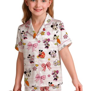 Puede incluir: Conjunto de pijama blanco con tem&aacute;tica de Pascua de Minnie Mouse. El conjunto incluye una camisa de manga corta con botones y pantalones cortos, ambos adornados con gr&aacute;ficos de Minnie Mouse, zanahorias, huevos y lazos rosas. El pijama tiene detalles de rayas rosas.