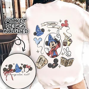 Op de afbeelding: Crèmekleurige sweatshirt met een kleurrijke afbeelding van Mickey Mouse als tovenaar, omringd door gerelateerde afbeeldingen. Bevat de tekst "Some imagination, huh?" en een zakontwerp.