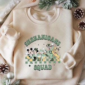 Puede incluir: Sudadera de color crema con un gráfico de personajes de Disney, incluyendo Mickey y Minnie Mouse, Donald y Daisy Duck, Goofy y Pluto. El texto "Shenanigans Squad" está impreso encima y debajo de los personajes en una fuente retro verde. El diseño está sobre un fondo a cuadros verde y blanco.