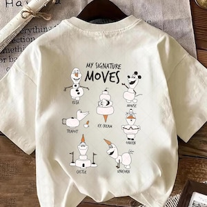 以下が含まれることがあります： 「MY SIGNATURE MOVES」の文字と、エルサ、ネズミ、ユニコーンなどのキャラクターの漫画イラストが描かれたクリーム色のTシャツ。クルーネックと半袖です。