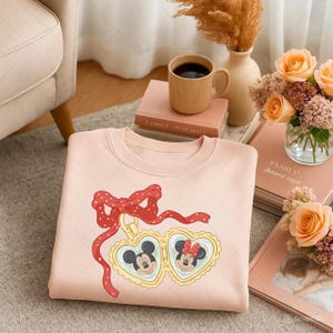 Op de afbeelding: Een lichtroze sweatshirt met een rode strik met witte stippen en een goudkleurig hartontwerp. Het hartontwerp bevat afbeeldingen van Mickey en Minnie Mouse. Het sweatshirt is op een lichtgekleurd oppervlak gevouwen.