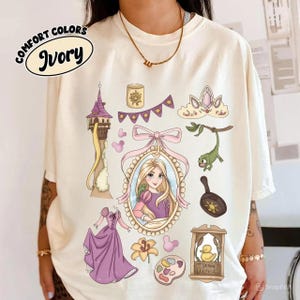 Op de afbeelding: Ivory Comfort Colors T-shirt met cartoon illustraties. Het ontwerp bevat een prinsessenportret, een toren, een kroon, een jurk en andere thema-elementen in paars, roze en geel. De tekst "Ivory" staat op het shirt.