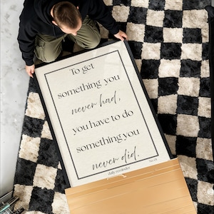 May include: A framed print with the text "To get something you never had, you have to do something you never did." The print is on a beige background with a black frame. The print is on a black and white checkered rug.