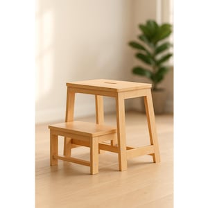 May include: A two-step wooden step stool in a light natural finish. The stool has a rectangular top with a small slot and a lower step. The legs are angled for stability. The stool is designed for reaching higher places.