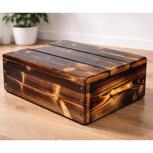May include: A rectangular, dark brown wooden storage box with a handle on the side. The box has a slatted top and is constructed with visible wood grain and metal fasteners. The wood has a charred appearance.