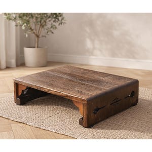 May include: A rectangular, wooden footstool with a dark brown finish. The footstool has a low profile with decorative cutouts on the sides and ornate legs. The surface has a textured wood grain pattern.