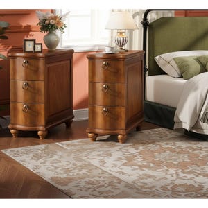 May include: Two wooden nightstands with three drawers each, featuring brass ring pulls. The nightstands are a warm brown color and sit on small, rounded feet. A bed with a green headboard is visible in the background.